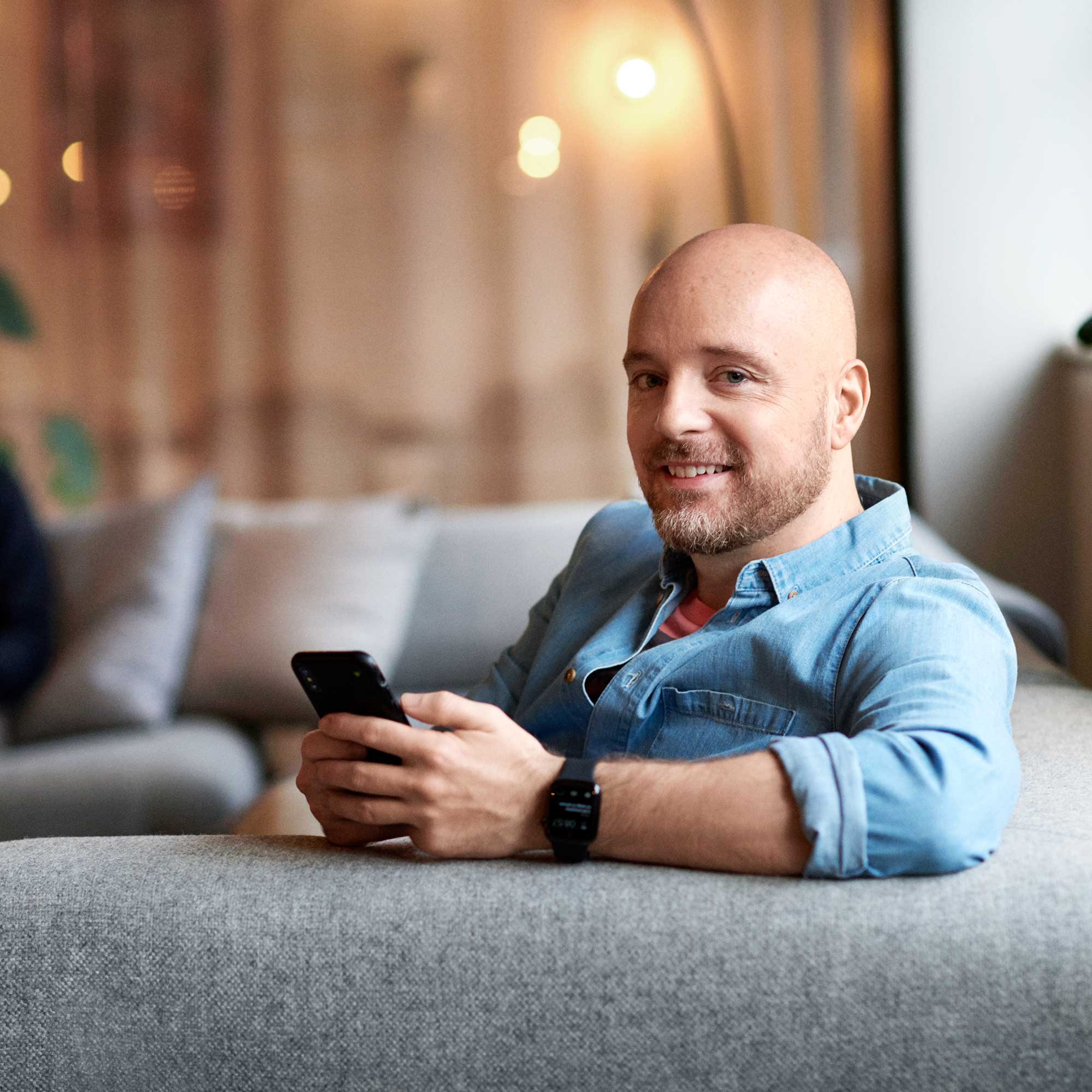 Leende man i jeansskjorta sitter i en grå soffa och håller i sin mobiltelefon